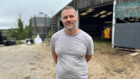 A man with short grey hair and a beard in a grey T shirt stands in front of a large wooden barn.