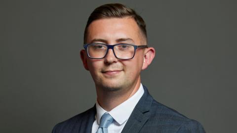 Parliamentary portrait of Crewe and Nantwich Labour MP Connor Naismith against a grey background. He has short, brushed-back brown hair, dark-rimmed glasses and is wearing a grey suit with pinstripe check, a white shirt and pale blue tie.