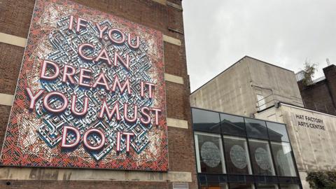 A large patterned design on a tall wall with the words IF YOU CAN DREAM IT YOU MUST DO IT in red and white lettering. There is a single-storey glass-fronted building to the right and a two storey concrete building to the right of that, labelled HAT FACTORY ARTS CENTRE.