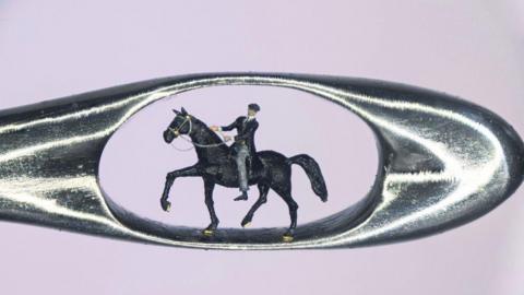 A micro sculpture of a man in a black coat, black hat and grey trousers sat in a horse inside the eye of a needle