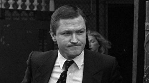 A black and white photo of Pat Finucane, with short dark hair. He is wearing a white shirt, dark tie and dark suit. 