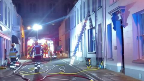 Two firefighters walking towards a fire engine with its doors open. On the right there is a ladder going up to a window. 