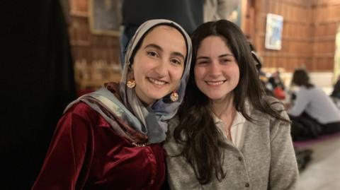Raza, who is wearing a patterned headscarf and a red-velvet top, sits next to Maya, who is wearing a greyish cardigan. They are looking at the camera and smiling.