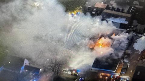 Aerial view of the fire with flames visible in the centre of an industrial building and smoke billowing from it. There are other buildings around the site. A fire engine is visible with blue lights.