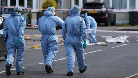 Three people in blue overalls, hats and masks on their way to a crime scene. A fourth person dressed the same is already at the scene, fiddling with something on the ground.