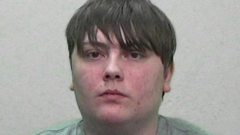 Mugshot of Elliot Robinson. He is clean shaven with a large round spotty face and greasy-looking brown hair, with a fringe below his eyebrows.