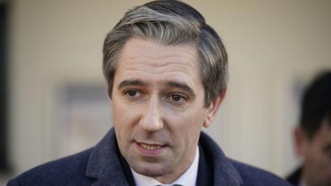 Simon Harris is mid speech looking to the side and the background is blurred. He has white grey hair and brown eyes. He is wearing a dark navy coat. 