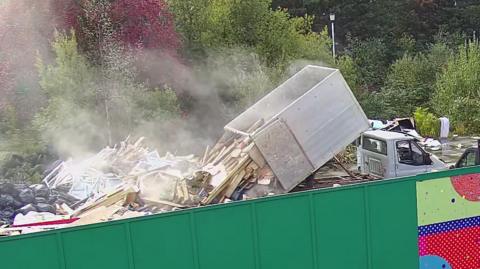 A tipper truck dumping bulky waste onto a mountain of existing rubbish. There is a green wall in the foreground and lots of trees in the background.