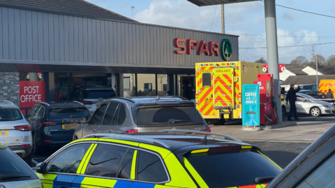 A wide shot of a petrol station forecourt, in the middle distance is a shop with SPAR in red letters and the green fir tree logo of the shop. An ambulance is parked outside on the right of the image, to its left a grey car can be seen in the shop window, behind it are other vehicles which are black and grey. In the foreground is a police car.