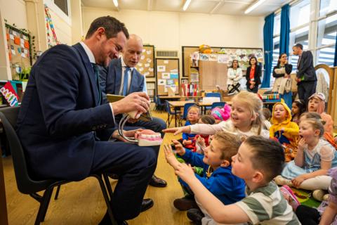 Children sit on the floor of a classroom learning how to brush their teeth. The health minister is sat in front of them with a large model of a mouth with bright white teeth and pink gums. The children in the first few rows are leaning forward to touch the model.