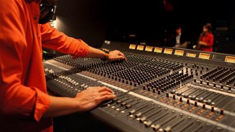 Man in orange shirt twisting black knobs on a grey recording desk