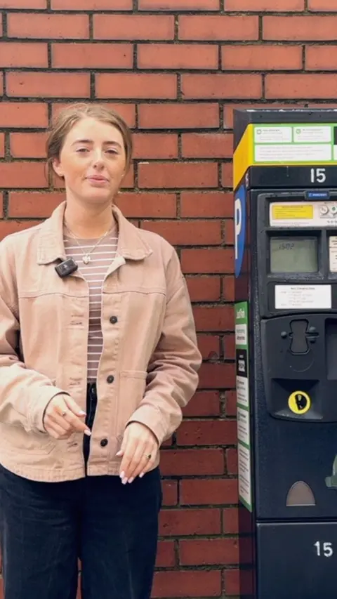 A woman with red hair wearing a pink jacket and black trousers standing in front of a red brick wall beside a black pay and display machine. 