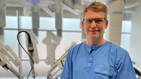 Gijs van Boxel, Consultant Upper GI Surgeon at Queen Alexandra Hospital is smiling and wearing light blue scrubs. He has glasses and short blonde hair. In the background four prongs of the da Vinci system.