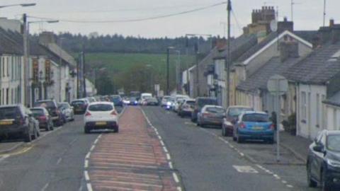 Killyleagh Street lined with houses and cars parked on either side. A grassy hill with trees atop is in the background. 