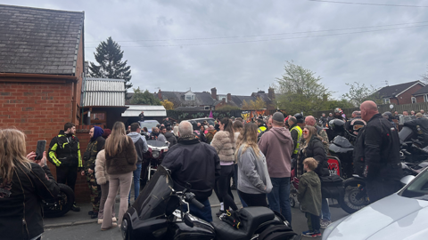 Hundreds of people outside a crematorium, with motorbikes in hte foreground