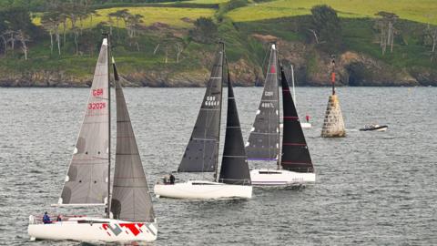 Three sailboats with sleek designs and modern sails on the water. Two have  black sails and the third, in the foreground, has grey. In the background, there is a stone beacon in the water, topped with a black and orange cone. A small motorboat is also on the water. The shoreline in the background is green with rolling hills and sparse trees.