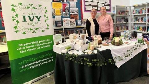 Two women manning a charity stand in a library with a banner reading The Ivy Trust supporting women who have experienced the death of a baby