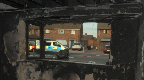 The view out of the window of a property, where the glass is missing from the window, and the walls are burnt. Emergency service vehicles can be seen in the road.