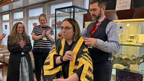 Lee Paton putting the new robe on the Mayor of Carlisle Jeanette Whalen. The new robe is black with intricate gold patterns. Onlookers are clapping.