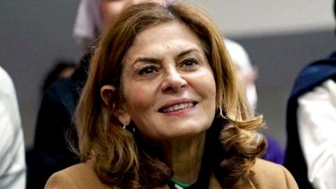 Close-up of Hind Kabawat, smiling. She has shoulder length brown hair and is wearing a smart, light brown coat.