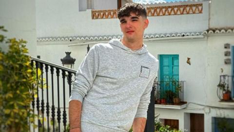 A photo of Joshua McBride, pictured with black hair and dark eyes. He is wearing a grey hoodie in the photo, taken in front of a balcony.