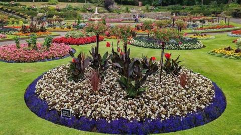 A floral display in a park has circular beds planted with rings on the outside and ornamental planting