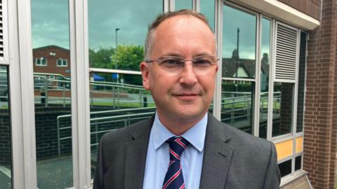 A man wearing a grey suit jacket, blue shirt and red and blue tie, stood outside a brick building. He is councillor Jonathan Lester, the Conservative leader of Herefordshire Council. 