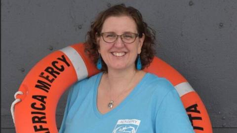 A photo of a woman called Ruth Cooper. She is in the middle of the photo and has short brown hair and dark rimmed glasses. She is wearing blue dangly earrings and a light blue top. Around her left shoulder is an orange life ring with Africa Mercy written in black on it.