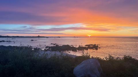 Vibrant sunset over calm water with purple, pink, and orange clouds stretching across the sky. The sun sits low on the horizon, illuminating the water dotted with rocks and small boats, while silhouetted coastal vegetation and a large boulder frame the foreground.