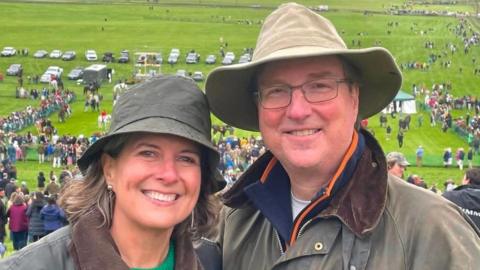 Alice Dearing and her husband John stood next to each other having their picture taken outside. They are dressed in coats and hats each and are looking directly at the camera and smiling. Alice is on the left and John is on the right. They are at an event with crowds of people in the background.