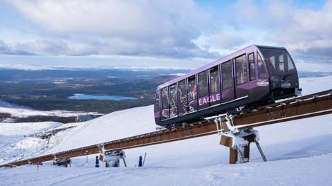 The funicular railway is a long metal rail installed on a mountainside. A purple, railway carriage-like vehicle travels along the rail. The ground below is covered in snow and beyond is sweeping landscape of hills and lochs.