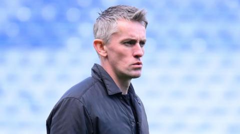 Side on view of ipswich town manger kieran mckenna who looks ahead with a focused look on his face and his lips slightly pouted. he wears a navy light jacket and stands in front of blue stadium seats.