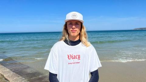 Jesse Heddercott is 16-year-old boy with long, blond hair past his shoulders. He is standing on a slipway. He wears a white cap, and a white T-shirt over a wetsuit that says Jersey Surf School. The sea is covering some of the slip, and is turquoise in the shallows before turning darker further out. The sky is blue.