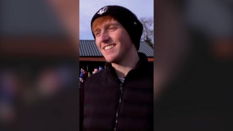 A man, Angry Ginge, smiling outside wearing a beanie hat and black jacket.