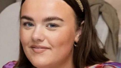 A close-up of Caitlin McLaughlin smiling at the camera. She has a headband in her brown hair.