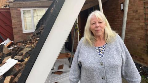 A woman stands in front of a fallen roof and pile of rubble. She is wearing a flowery top and grey cardigan and has long blond hair. The rubble is in front of part of her home which has a window and brick wall in. 