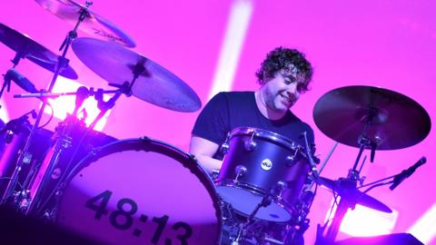 Ian Matthews playing the drums at a Kasabian gig. He is looking down whilst he plays and is smiling. The stage is lit in pink lighting.