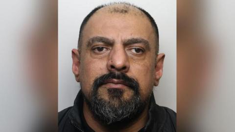 Police custody image of Suaiel Haider. He has receding black hair and a beard and is wearing a black shirt.