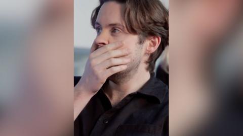 A man with short, brown hair covers his mouth with his hand