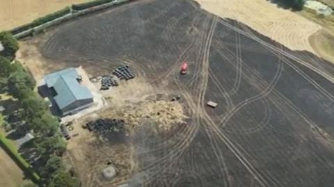 A blackened field has tracks over it and smouldering ground. Therer are trees and buildings close to where the blaze was. 