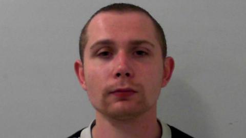 Mugshot of a man with a short goatee and a buzzcut. He has a neutral expression. He is wearing a grey jumper.