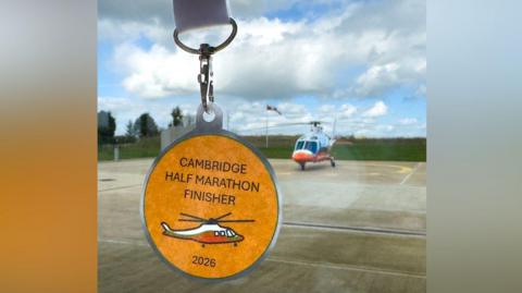A medal is at the forefront on the left and is orange. It has Cambridge Half Marathon Finisher 2026 written on it and a picture of an air ambulance. In the background is a white and orange air ambulance which is sat on a helipad.