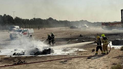 Firefighters work at the scene of a plane crash, water spraying onto a burnt piece of metal on the ground, it is in an open stretch of land with cars and a fence visible in the background.