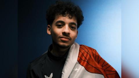 A young man with black hair is looking at the camera, he is wearing a black jacket and has the Welsh flag draped on his left shoulder. The background is a wall that is a mix of light and dark blue.