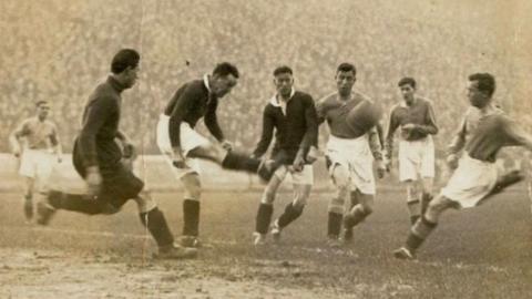 A black and white old photograph of a football match. Three players in black are competing for the ball with three other players in lighter-coloured kits.