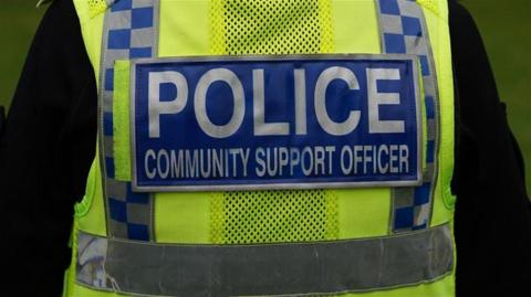 Picture of the back of a Police Community Support Officer in uniform with a hi vis police jacket on. The jacket has white writing on a blue background with the word police in bold. 