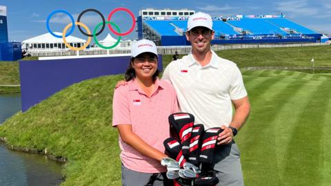 Shannon Tan and her caddie Robin Ellis, practicing on Le Golf National course at the Paris Olymipcs 2024. Shannon is on the left wearing a pink t-shirt and white cap, Robin is on the right wearing a white t-shirt and white cap, and they are holding golf clubs in front of them