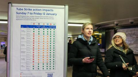 An image of a poster with information about the Tube strike action. Two women walk past the sign wearing long black puffer jackets and one wearing a wooly hat. 