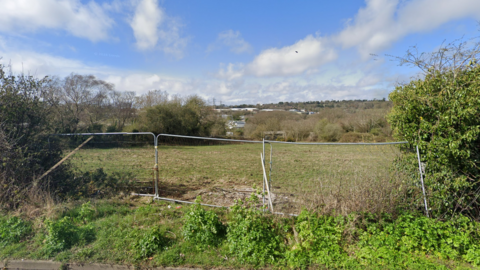 A view of a grass field through a gap in the hedgerow looking out over Hastings. There are three temporary metal fences blocking the entrance. The sky is blue and cloudy