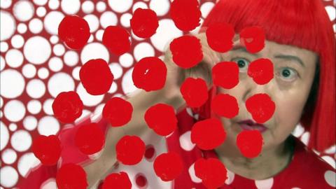 Yayoi Kusama seen looking through a clear screen with red spots painted on it, with red and white clothes and background